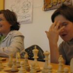 Zwei Kinder sitzen an einem Tisch und spielen Schach; eines lächelt, während das andere einen Teil seines Gesichts mit einer Hand verdeckt. Die Schachfiguren sind auf dem Brett vor ihnen angeordnet.