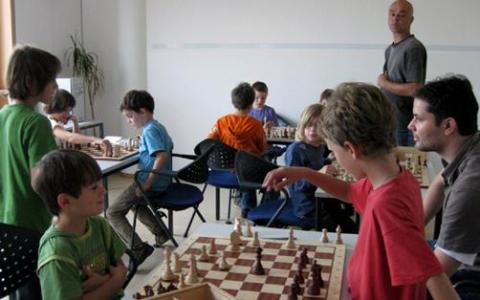 Kinder und Erwachsene spielen Schach an Tischen in einem Klassenzimmer, wobei ein Erwachsener den Raum beobachtet.
