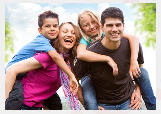 Zwei Erwachsene und zwei Kinder befinden sich im Freien, wobei jedes Kind von einem Erwachsenen auf dem Rücken getragen wird. Alle vier lächeln in die Kamera.