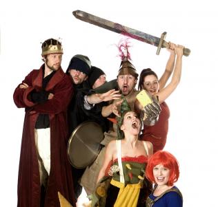 Sechs Erwachsene in verschiedenen mittelalterlichen oder Fantasiekostümen posieren spielerisch; einer trägt eine Krone, ein anderer hält ein Schwert über die Gruppe, andere halten Requisiten und machen ausdrucksvolle Gesichter.
