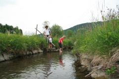 Zwei Kinder und ein Erwachsener stehen an einem kleinen Bach, die Kinder waten im Wasser, und auf beiden Seiten gibt es grasbewachsene Ufer.