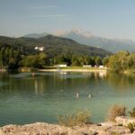 Ein ruhiger See mit zwei Schwimmern im Wasser, umgeben von Bäumen und Bergen unter einem klaren Himmel. Am Ufer sind Gebäude und in der Ferne ein Schloss zu sehen.