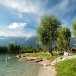 Menschen entspannen sich an einem Seeufer mit einem kleinen Boot auf dem Kiesstrand, Bäumen als Schattenspender und Bergen im Hintergrund unter einem teilweise bewölkten Himmel.