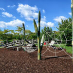 Spielplatz im Freien mit Klettergerüsten aus Holz, einem Seilnetz und grünen Bäumen unter einem blauen Himmel mit vereinzelten Wolken.