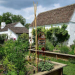 Ein Garten mit Hochbeeten mit verschiedenen Pflanzen vor zwei weißen Gebäuden im Landhausstil unter einem teilweise bewölkten Himmel.