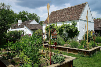 Ein Garten mit Hochbeeten mit verschiedenen Pflanzen vor zwei weißen Gebäuden im Landhausstil unter einem teilweise bewölkten Himmel.
