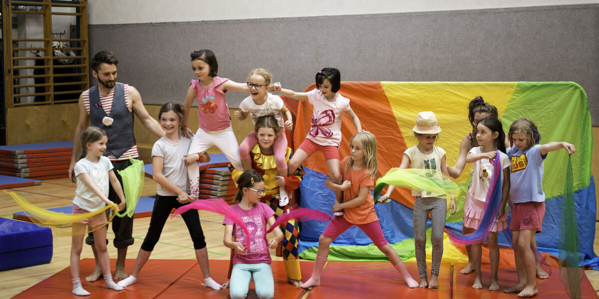 Eine Gruppe von Kindern und ein Erwachsener posieren auf Turnmatten in einer Halle, einige halten bunte Tücher in der Hand, und im Hintergrund ist ein Regenbogen zu sehen.