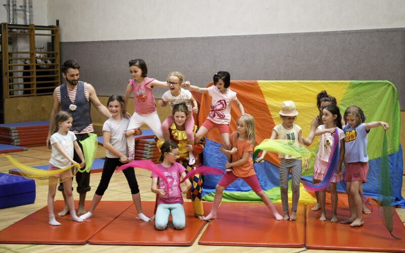 Eine Gruppe von Kindern und ein Erwachsener posieren auf Turnmatten in einer Halle, einige halten bunte Tücher in der Hand, und im Hintergrund ist ein Regenbogen zu sehen.