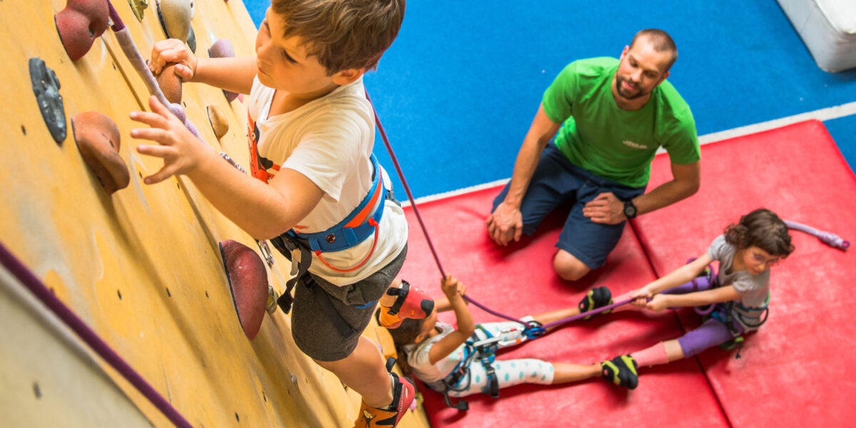 Ein kleiner Junge klettert an einer Indoor-Felswand, während ein Mann die Aufsicht führt und zwei Kinder auf Matten mit Kletterseilen umgehen.
