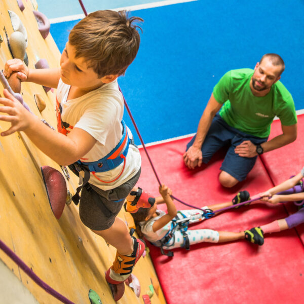 Ein kleiner Junge klettert an einer Indoor-Felswand, während ein Mann die Aufsicht führt und zwei Kinder auf Matten mit Kletterseilen umgehen.