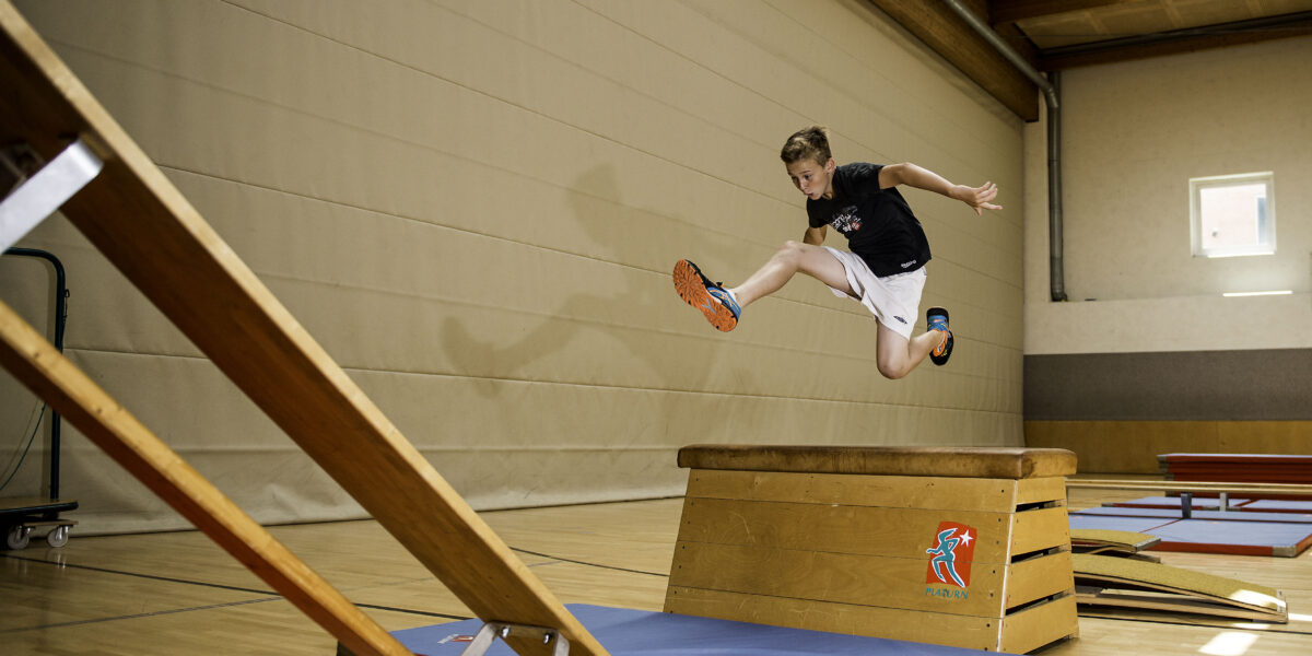 Ein Junge in Sportkleidung springt über einen hölzernen Turnkasten auf eine Matte in einer Sporthalle.