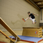 Ein Junge in Sportkleidung springt über einen hölzernen Turnkasten auf eine Matte in einer Sporthalle.