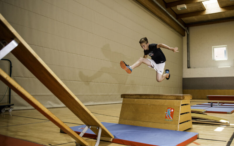 Ein Junge in Sportkleidung springt über einen hölzernen Turnkasten auf eine Matte in einer Sporthalle.