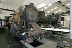 Eine verrostete, ausgemusterte Dampflokomotive steht in einem Innenraum auf einem Gleisabschnitt, während im Hintergrund ein anderer Zug zu sehen ist.