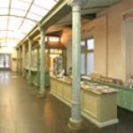 Ein Flur mit hohen Fenstern, Säulen und Tischen, auf denen verschiedene Gegenstände ausgestellt sind, wahrscheinlich in einem Museum oder einer Galerie mit Geschenkartikeln.