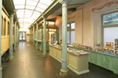 Ein Flur mit hohen Fenstern, Säulen und Tischen, auf denen verschiedene Gegenstände ausgestellt sind, wahrscheinlich in einem Museum oder einer Galerie mit Geschenkartikeln.