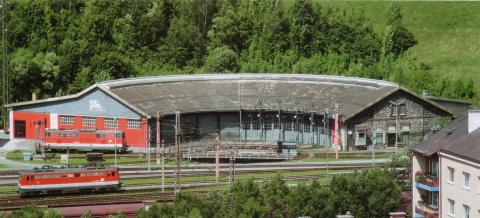 Ein großer, halbkreisförmiger Ringlokschuppen mit mehreren Türen, umgeben von Gleisen und Grünanlagen, vor dem eine rote Lokomotive zu sehen ist.