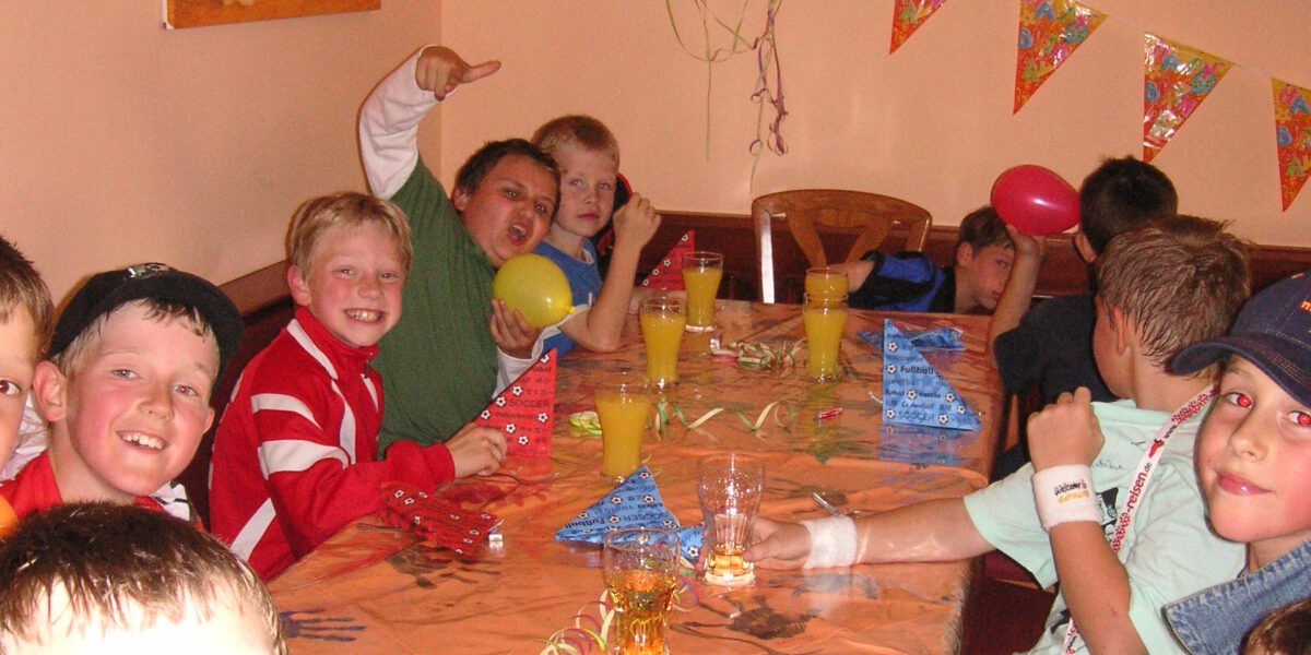 Eine Gruppe von Kindern sitzt bei einer Party um einen geschmückten Tisch mit Getränken und Luftballons; im Hintergrund sind Papierhüte und ein Fernseher zu sehen.