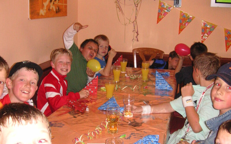 Eine Gruppe von Kindern sitzt bei einer Party um einen geschmückten Tisch mit Getränken und Luftballons; im Hintergrund sind Papierhüte und ein Fernseher zu sehen.
