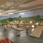 Indoor-Minigolfplatz mit mehreren Putting-Greens und Hindernissen, dekoriert mit Pflanzen, Laternen und einem Wandgemälde mit einer malerischen Landschaft an der Rückwand.
