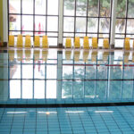 Ein Hallenbad mit klarem Wasser, blauen Kacheln und einer Reihe gelber Stühle vor großen Fenstern, die das Tageslicht hereinlassen.