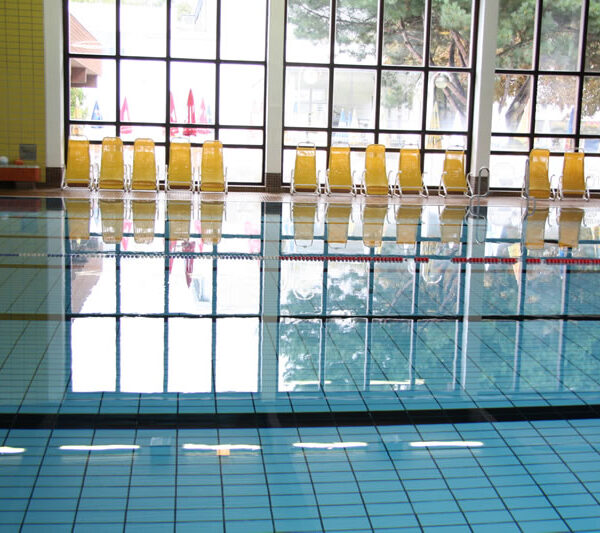 Ein Hallenbad mit klarem Wasser, blauen Kacheln und einer Reihe gelber Stühle vor großen Fenstern, die das Tageslicht hereinlassen.