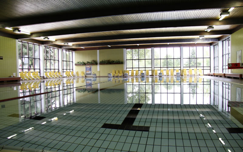 Hallenbad mit ruhigem Wasser, gefliestem Boden, großen Fenstern und Reihen von gelben Stühlen an den Wänden.