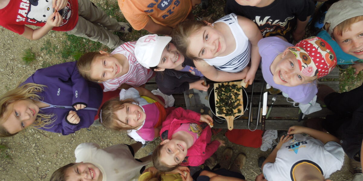 Eine Gruppe von Kindern steht im Freien in einem Kreis und blickt in die Kamera, in der Mitte steht eine Schale mit Essen auf einem Holztisch.