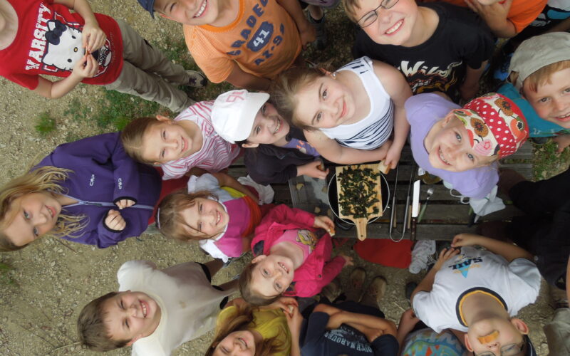 Eine Gruppe von Kindern steht im Freien in einem Kreis und blickt in die Kamera, in der Mitte steht eine Schale mit Essen auf einem Holztisch.