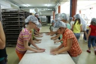 Eine Gruppe von Personen mit Haarnetzen arbeitet an einem langen Tisch in einer Großküche oder Bäckerei.