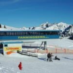 Eine Skiliftstation mit der Aufschrift "Fanningberg" steht an einem verschneiten Berghang, in der Nähe befinden sich mehrere Personen in Winterkleidung und im Hintergrund sind schneebedeckte Gipfel zu sehen.