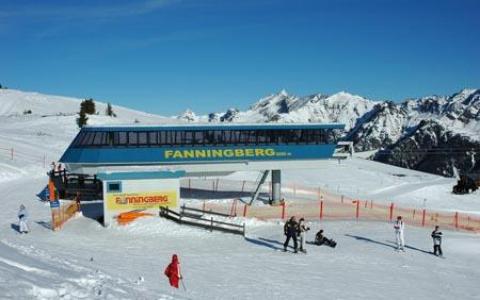Eine Skiliftstation mit der Aufschrift "Fanningberg" steht an einem verschneiten Berghang, in der Nähe befinden sich mehrere Personen in Winterkleidung und im Hintergrund sind schneebedeckte Gipfel zu sehen.