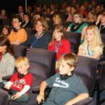 Eine Gruppe von Erwachsenen und Kindern sitzt in einem Hörsaal und sieht sich eine Veranstaltung oder Präsentation an, wobei einige von ihnen Kopfhörer tragen.