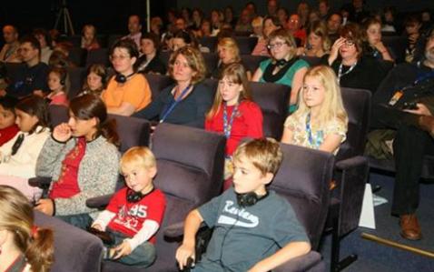 Eine Gruppe von Erwachsenen und Kindern sitzt in einem Hörsaal und sieht sich eine Veranstaltung oder Präsentation an, wobei einige von ihnen Kopfhörer tragen.