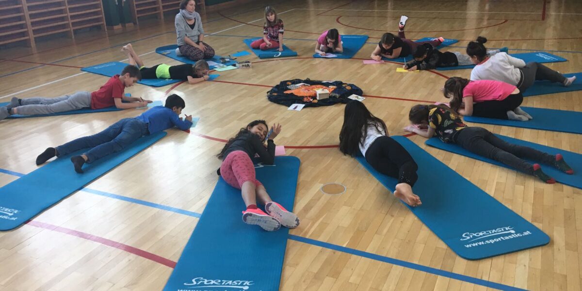 Kinder liegen auf Yogamatten in einem Kreis auf dem Boden einer Turnhalle, ein Erwachsener sitzt in der Nähe und in der Mitte liegen Papiere oder Karten.