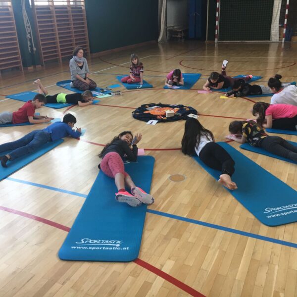 Kinder liegen auf Yogamatten in einem Kreis auf dem Boden einer Turnhalle, ein Erwachsener sitzt in der Nähe und in der Mitte liegen Papiere oder Karten.