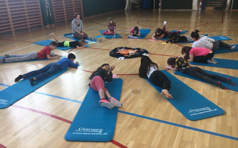 Kinder liegen auf Yogamatten in einem Kreis auf dem Boden einer Turnhalle, ein Erwachsener sitzt in der Nähe und in der Mitte liegen Papiere oder Karten.