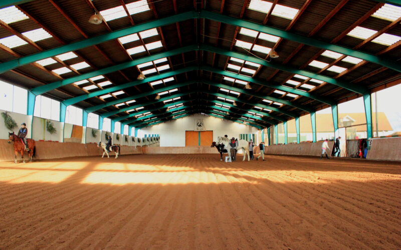 Reithalle mit mehreren Personen und Pferden, sandigem Boden, hoher Decke und Oberlichtern, die für natürliche Beleuchtung sorgen.