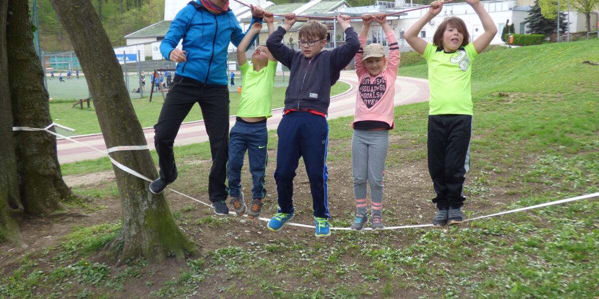 Fünf Kinder balancieren auf einer Slackline zwischen Bäumen und halten sich dabei an einem Seil fest.