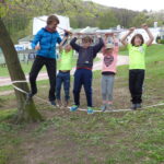 Fünf Kinder balancieren auf einer Slackline zwischen Bäumen und halten sich dabei an einem Seil fest.