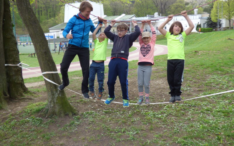 Fünf Kinder balancieren auf einer Slackline zwischen Bäumen und halten sich dabei an einem Seil fest.