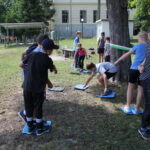 Eine Gruppe von Kindern nimmt an einer Aktivität im Freien teil. Sie stehen auf Matten und strecken sich einander auf einer Wiese in der Nähe von Bäumen und einem Gebäude entgegen.