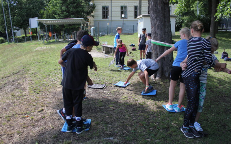 Eine Gruppe von Kindern nimmt an einer Aktivität im Freien teil. Sie stehen auf Matten und strecken sich einander auf einer Wiese in der Nähe von Bäumen und einem Gebäude entgegen.