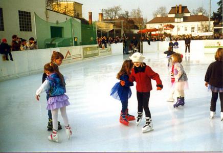 Kinder in farbenfrohen Kostümen beim Schlittschuhlaufen auf einer Freiluft-Eisbahn, während die Zuschauer hinter der Absperrung zuschauen.