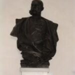 Bronzebüste eines Mannes mit Schnurrbart, der eine Jacke trägt und auf einem weißen Sockel vor einer schlichten Wand steht.