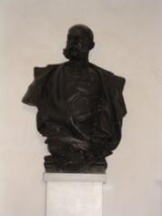 Bronzebüste eines Mannes mit Schnurrbart, der eine Jacke trägt und auf einem weißen Sockel vor einer schlichten Wand steht.