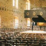 Ein Flügel steht auf einer Bühne in einem leeren Konzertsaal mit Steinwänden, Holzstühlen und großen Fenstern.