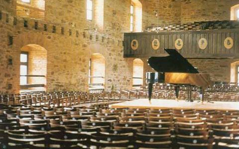 Ein Flügel steht auf einer Bühne in einem leeren Konzertsaal mit Steinwänden, Holzstühlen und großen Fenstern.