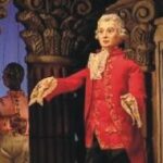 Eine Puppe in einem verzierten roten Mantel mit weißer Spitze steht vor detaillierten architektonischen Säulen.