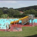 Öffentliches Freibad mit Menschen, die schwimmen und sich entspannen; eine gelbe Rutsche teilt das Becken, mit Grasflächen und Bäumen im Hintergrund.